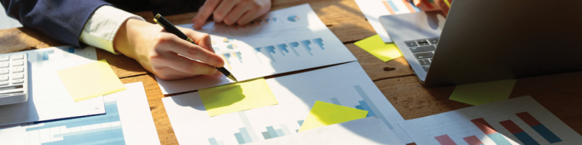 hand holding a pen writing on yellow sticky notes with graphs on papers underneath and a computer on the table