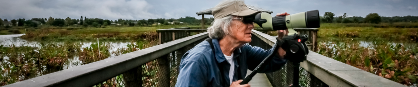 older woman with gray hair in blue shirt looking through a pair of binoculars outside