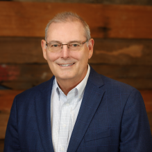 man in light shirt with dark blazer smiling against wood wall