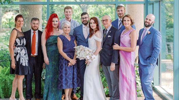 11 people stand in front of window smiling. Bridge has wedding dress and flowers.