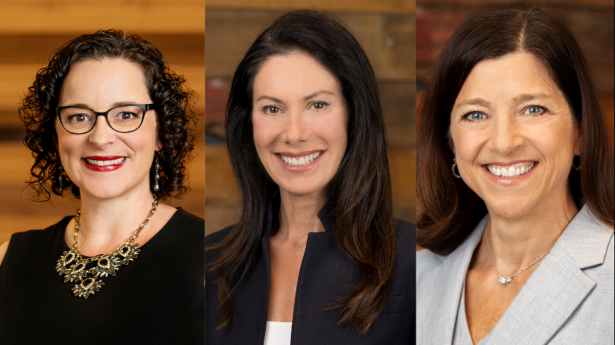 Three women all smile at camera side by side. Woman on left has black dress on and necklace over dress. Woman in center has a dark suit on with dark brown hair. Woman on right has dark brown hair and grey suit on.