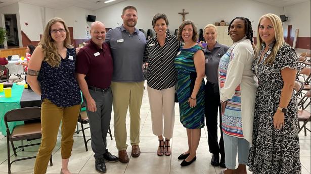 Eight people stand next to each other smiling with cross hanging in background at a church facility.