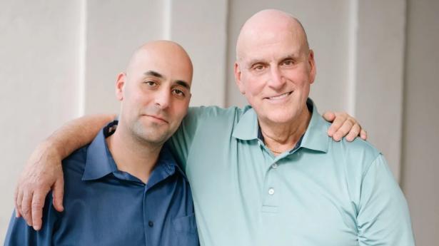 Father-son duo hold arms around each other. Both have no hair and are wearing blue toned shirts.