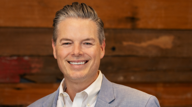 President | CEO of Gulf Coast Community Foundation, Phillip Lanham, wears grey suit jacket and white shirt with dark wood wall in background. Smiles.