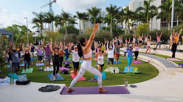 Woman stands at front of outdoor class holding yoga pose with arms up and students following.