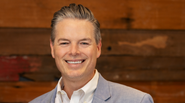 One man with grey suit jacket and white shirt smiles at camera with dark wood wall in background.