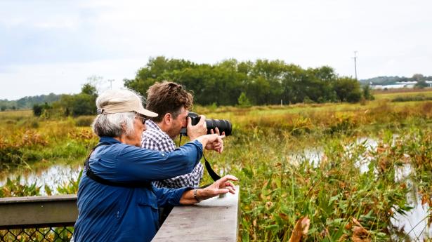 older woman and young man looking through a pair of binoculars and pointing out across a pond