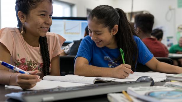 Two girls with dark brown hair smile, sitting at desk with notebooks and paper around them.