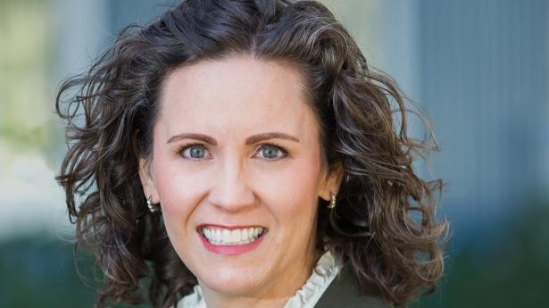 One woman with short, curly dark hair smiles at camera outdoors with grey suit on and white shirt. 