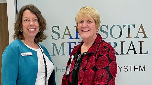 Two women stand smiling, one wearing teal jacket and one wearing red and black jacket with roses.