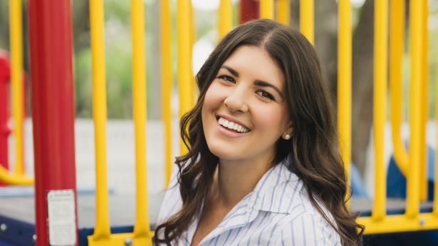 One woman with long brown hair and white striped shirt smiles at camera with yellow, red, and blue playground behind her.