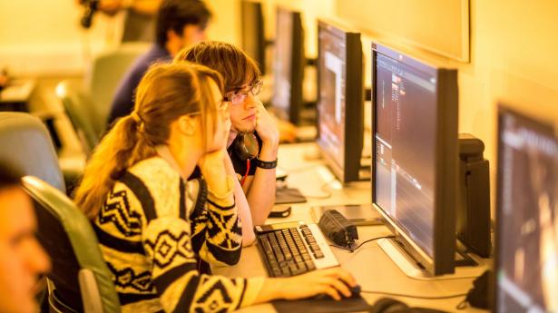 Two students sitting side by side looking at computer screen, one wearing sweater and one wearing watch and glasses.