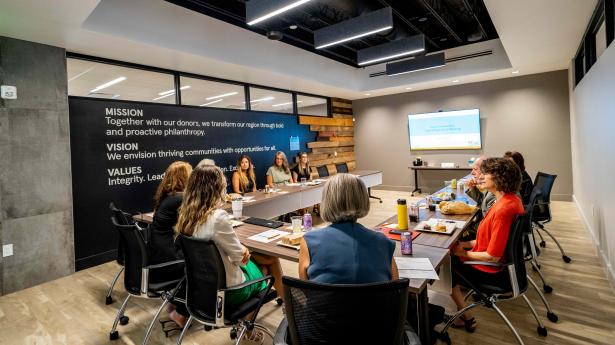 About 8 people sit at a table in a room with a black wall with mission, vision, and values on it and salvaged wood.