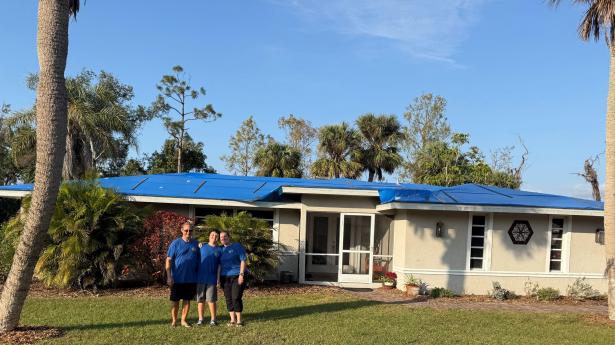 Three people stand outside home with blue tarp on roof.