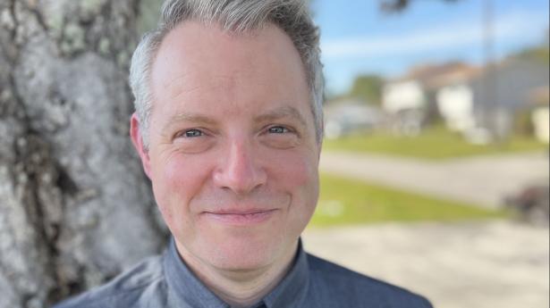 One man in blue shirt with grey hair smiles at camera near tree outside.