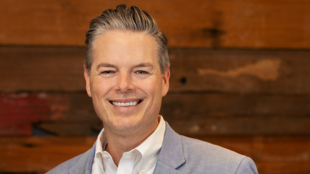 One man stands and smiles with grey suit jacket on, white shirt, and wood wall behind him.