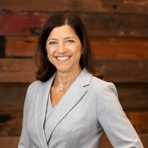 woman in a light blue suit smiling at the camera against a dark wooden wall