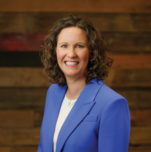 woman in a blue suit and white shirt with curly brown hair smiles at the camera against a wooden wall