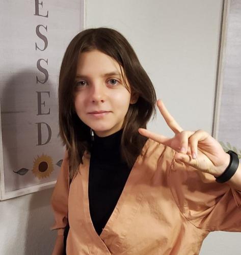 One young woman stands with tan suit jacket and black shirt holding peace symbol.