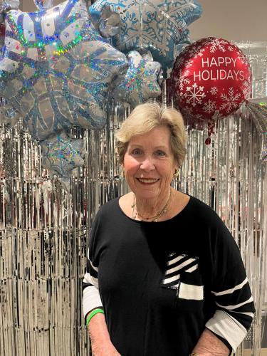 Woman stands around happy holidays balloons smiling with black and white sweater on.