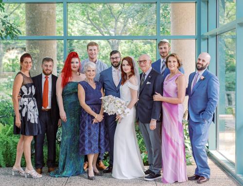 11 people smile at camera near window. Bridge is wearing wedding dress and holding flowers.