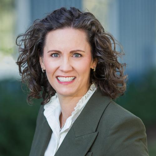 Woman smiles outdoors with dark, curly hair, grey suit jacket, and white shirt.