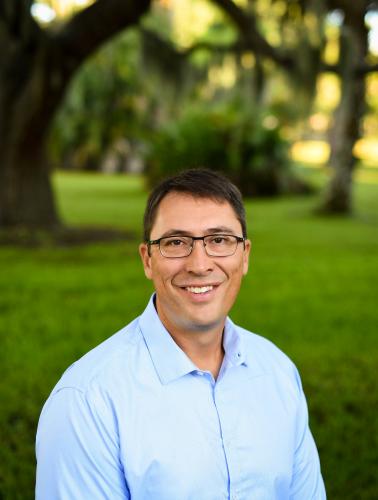 One man stands outside with glasses, brown hair, and blue shirt.