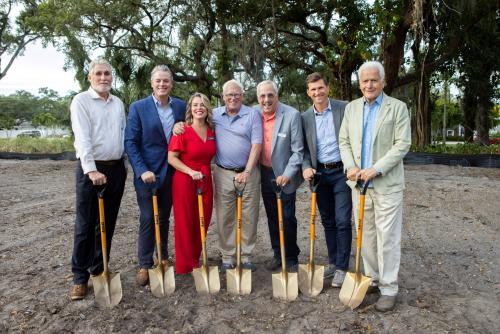 Seven people stand outside in dirt lots smiling with shovels from a veterans housing groundbreaking.