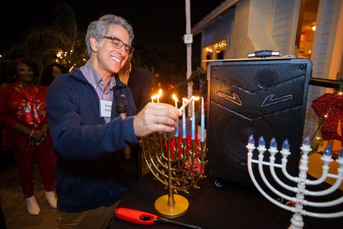 A rabbi stands outside in the dark lighting a gold menorah with blue candles. Another menorah sits closeby. A speaker is also on the table with the menorahs.