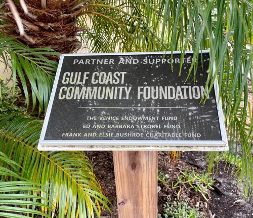 Black sign around trees sitting on wood poll. Sign says: Partner and Supporter, Gulf Coast Community Foundation, The Venice Endowment Fund, Ed and Barbara Strobel Fund, Frank and Elsie Bushroe Chiirtable Fund