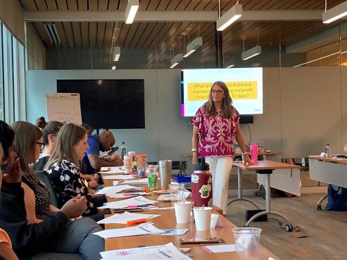 Woman stands in front of room with pink shirt. Participants at I3 workshop are gathered, sitting at tables around her.