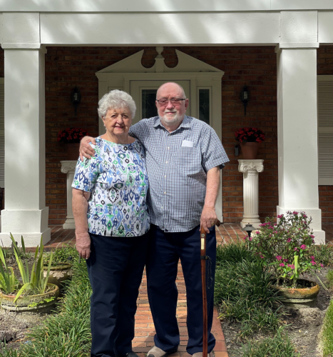 One man stands with cane and one woman stands, smiling at camera, holding one another.