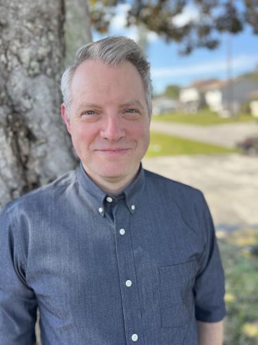 One man with grey hair and blue shirt stands outside near tree.