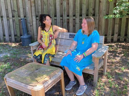 Two women sit outside on bench smiling. Wood fence is behind them.