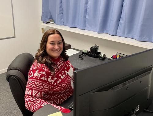 Woman wearing red sweater with brown hair looks up from computer and desk, smiling.