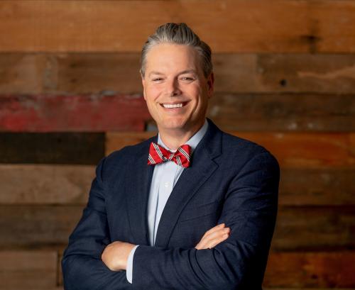 Man with suit and red striped bowtie smiles with wood wall in background.
