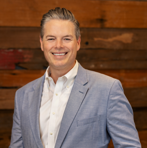 One man stands in front of dark wood wall with grey suit jacket, smiling.