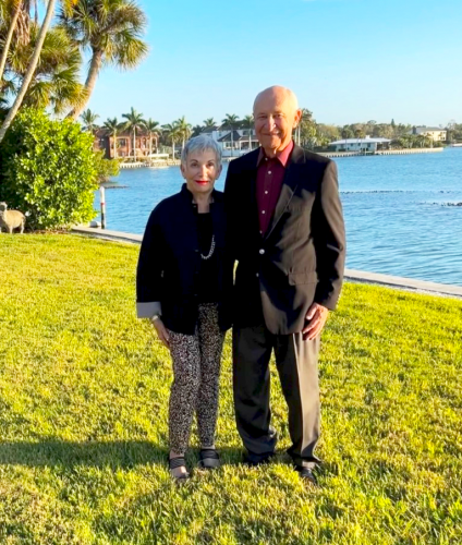 A man and a woman both in black jackets smile at camera with ocean view behind them.