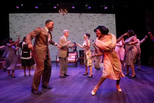 Two people dance on stage, one wearing orange dress. Other people dance behind them.