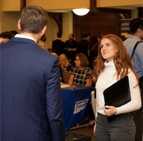 One young woman with long sleeved white shirt and long red hair holding black folder looks up in conversation.