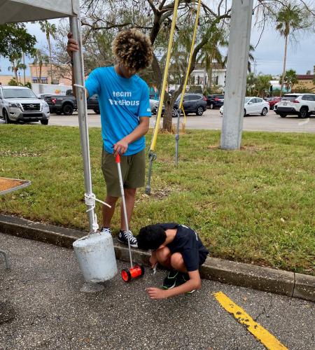 Two students stand outside by tent, one helping with something on the ground and the other standing.