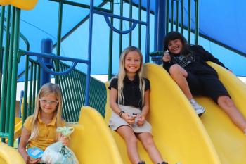 three children on the top of yellow slides smiling at the camera outside