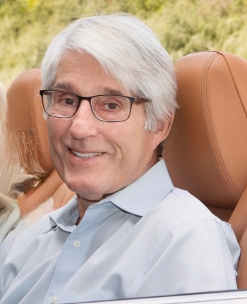 older man with glasses and white hair sitting in the front seat of a car smiling at the camera