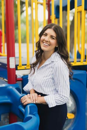 One young woman with long brown hair and striped shirt smiles with playground in background.