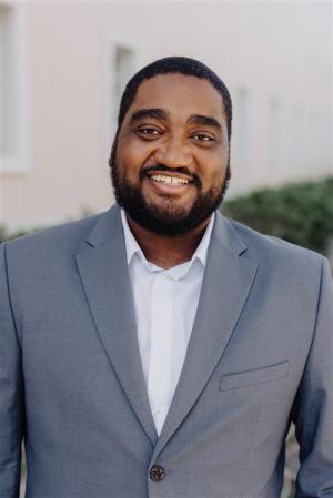 One African American man stands smiling in grey suit.